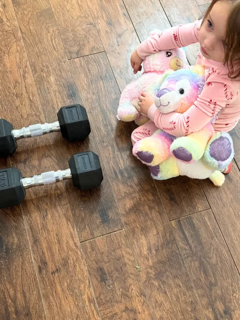 toddler sitting with stuffed animals next to dumbbells on living room floor